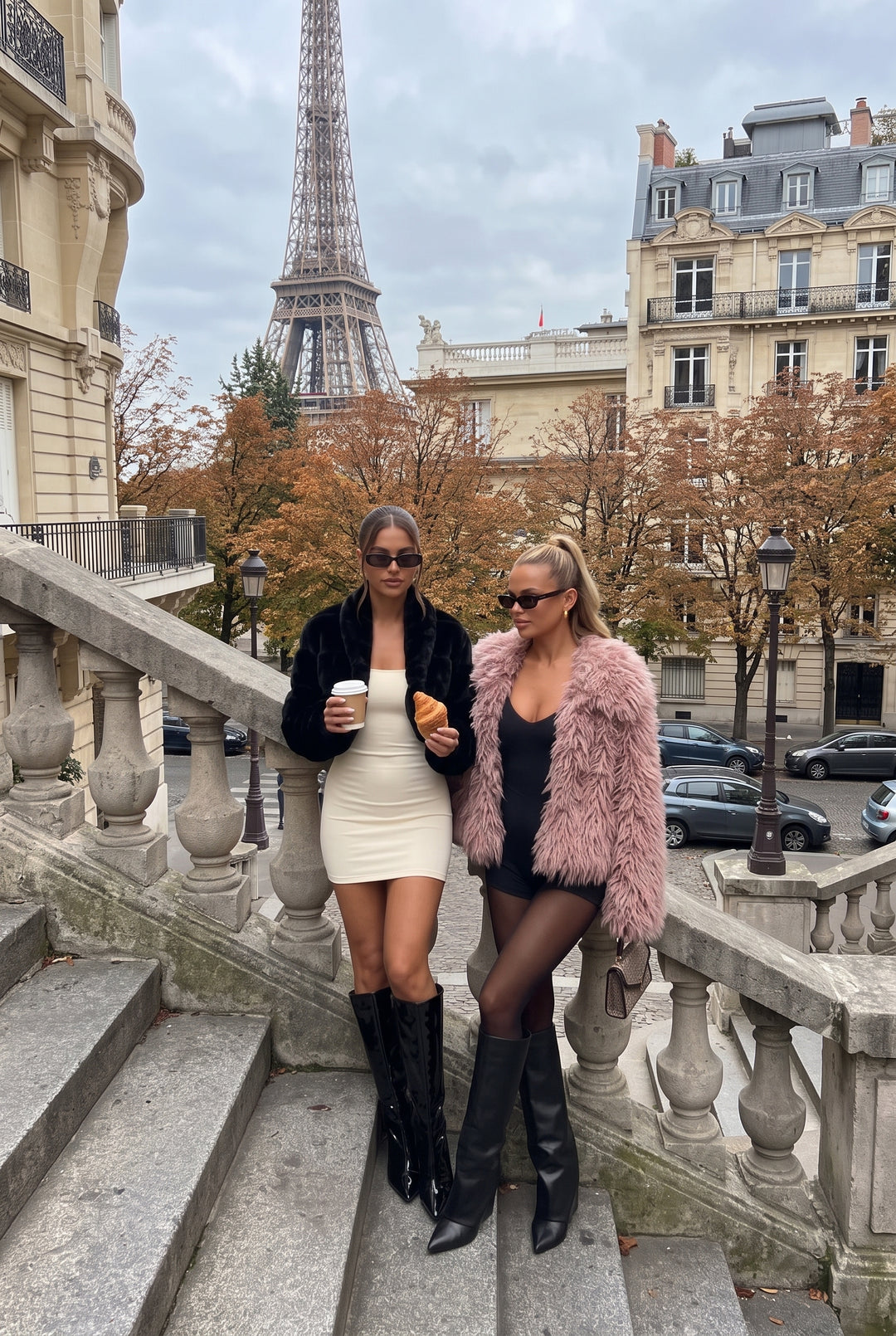 Two women posing on stone stairs in Paris with the Eiffel Tower and buildings in the background. One wears a black faux fur jacket over a cream mini dress and black knee-high boots, holding a coffee cup and a pastry.