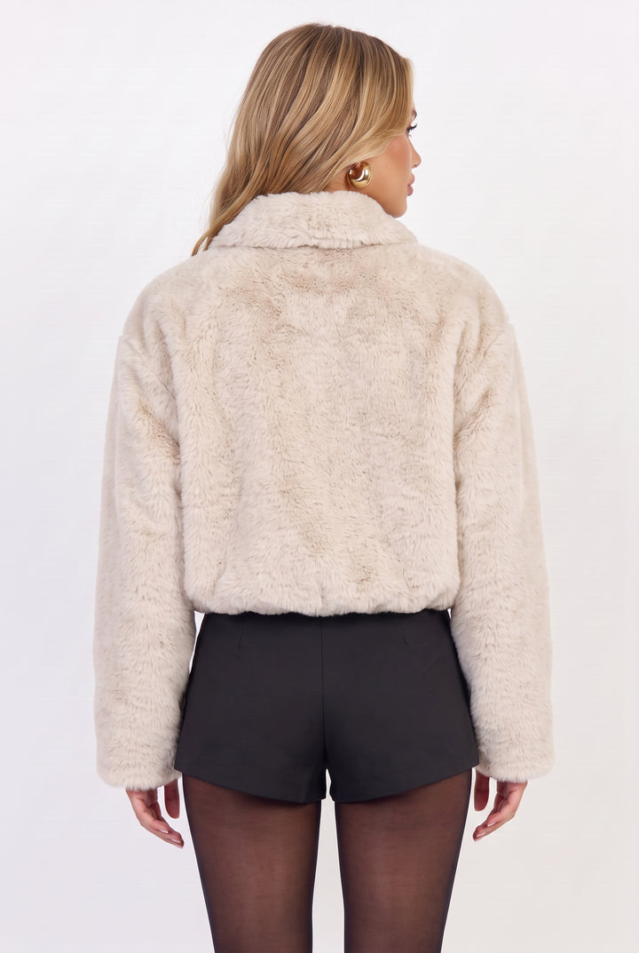 A woman shown from the back wearing a cropped, long-sleeve beige faux fur jacket with a high collar, paired with black shorts and sheer black tights, against a plain white studio background.