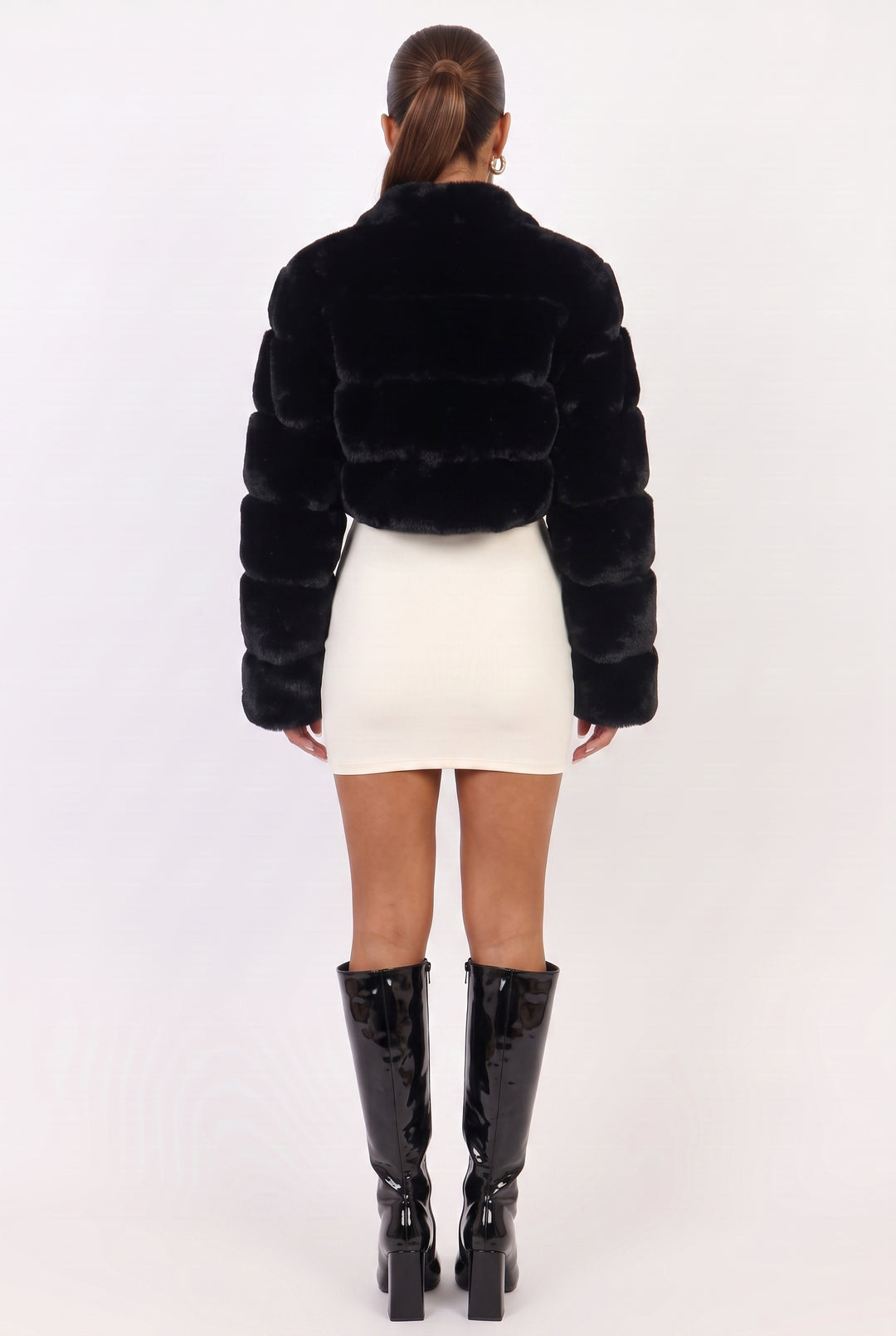 A model shown from the back wearing a black cropped faux fur jacket, a white mini skirt, and black knee-high patent leather boots against a plain white studio background.