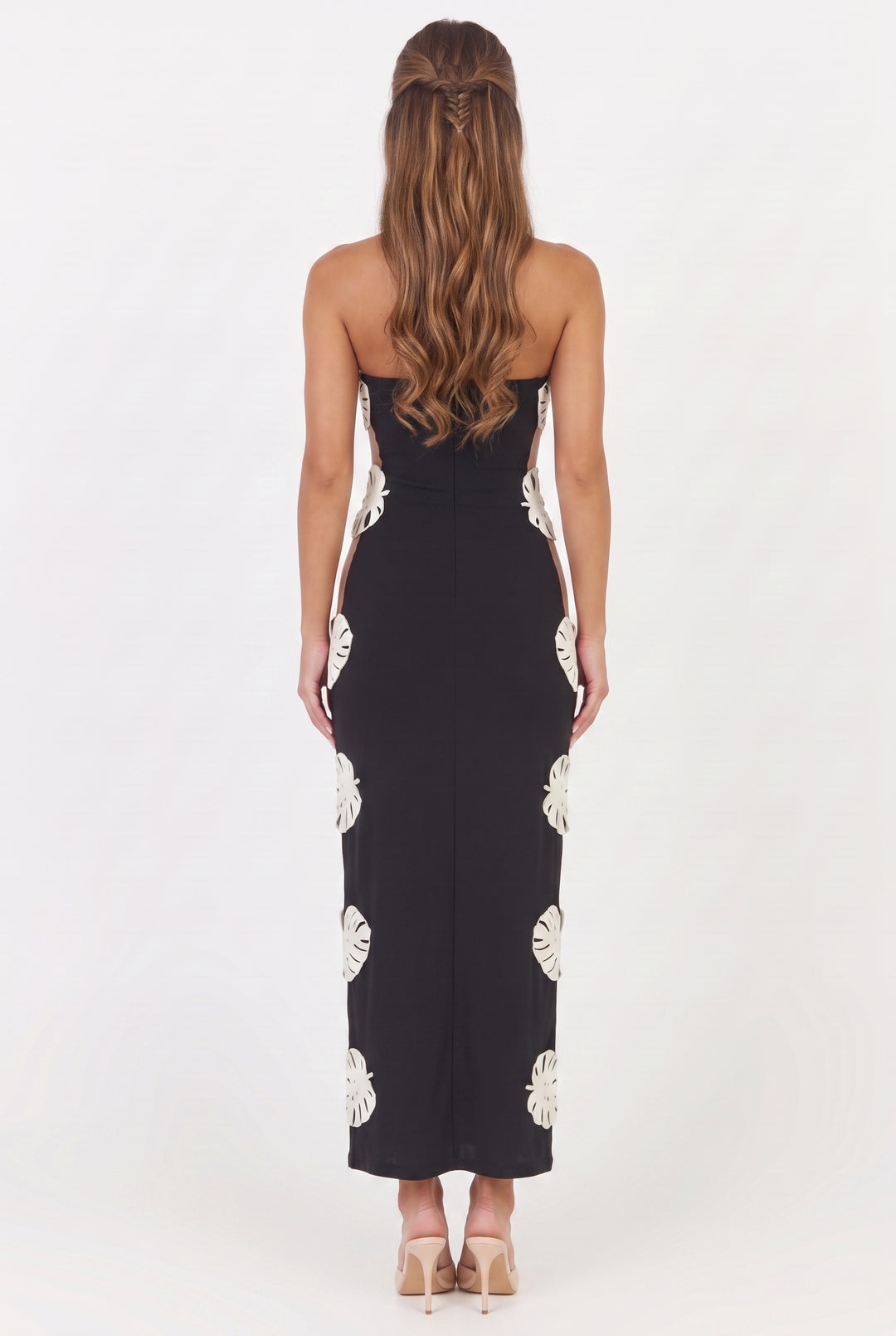 A woman viewed from the back wearing a strapless black maxi dress with large white monstera leaf prints down the sides, paired with beige high heels against a plain white studio background.