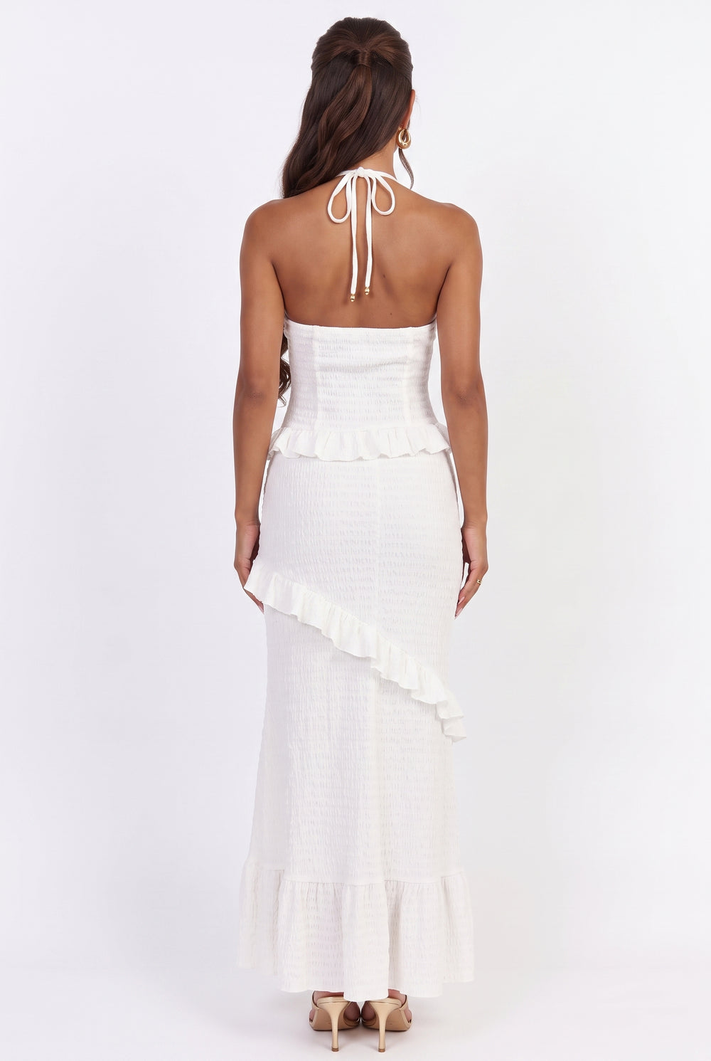 A woman viewed from the back wearing a white strapless halter maxi dress with a ruffled peplum and asymmetrical ruffle detail, paired with beige high heels against a plain white studio background.
