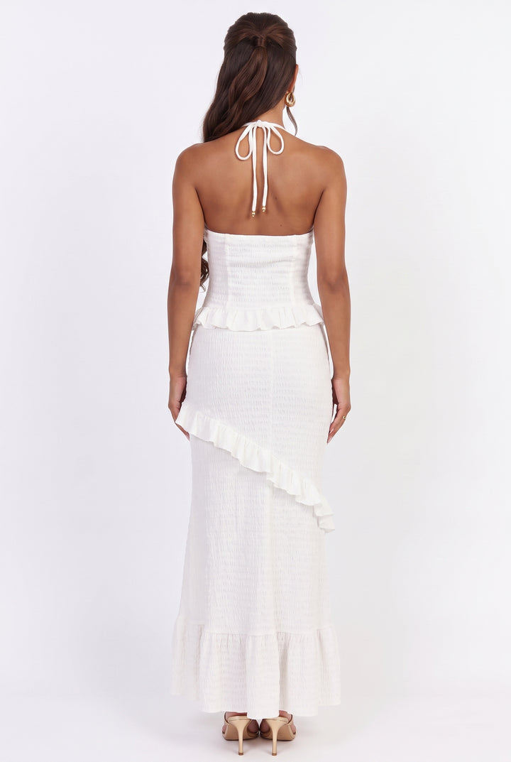 A woman viewed from the back wearing a white strapless halter maxi dress with a ruffled peplum and asymmetrical ruffle detail, paired with beige high heels against a plain white studio background.