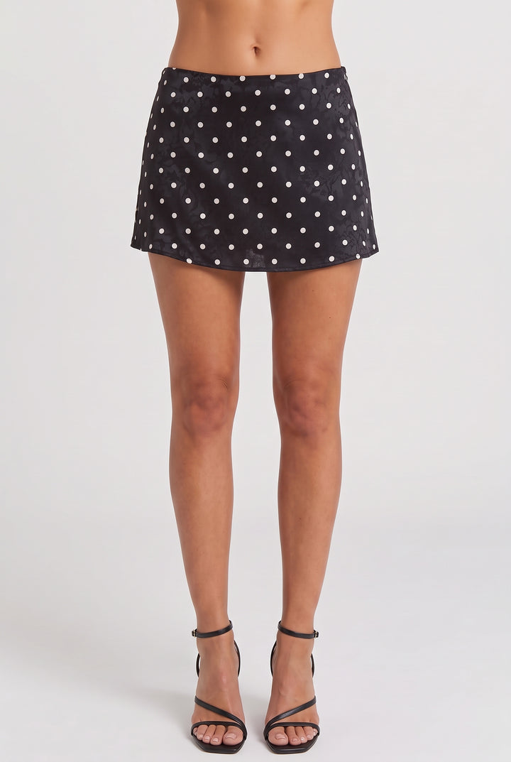 A model's lower body wearing a black mini skirt with a white polka dot pattern and black strappy high-heeled sandals, photographed against a light gray studio background.